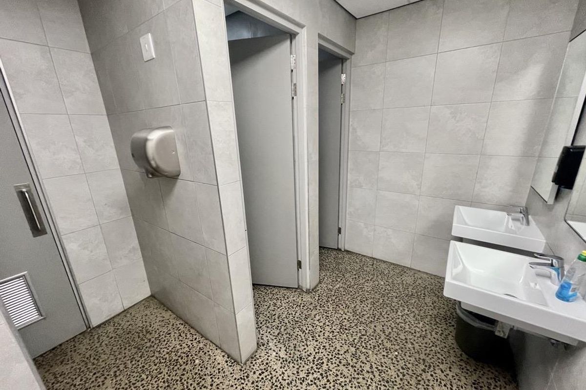 A clean industrial-style communal restroom featuring tiled walls, exposed aggregate flooring, dual wall-mounted basins, and multiple private cubicle entrances.