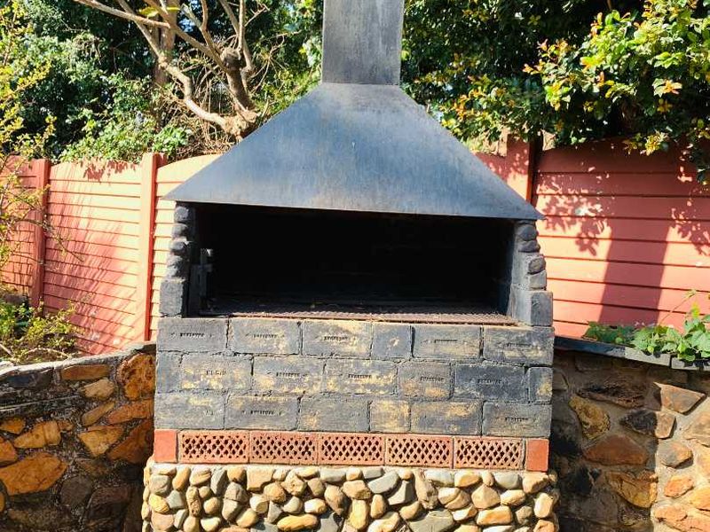 Outdoor patio area featuring a rustic built-in stone and brick braai with a metal chimney, set against a garden backdrop.