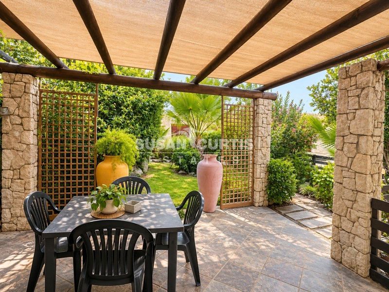 A covered outdoor patio featuring stone-clad pillars, a timber pergola with shade cloth, and tiled flooring, overlooking a landscaped garden.
