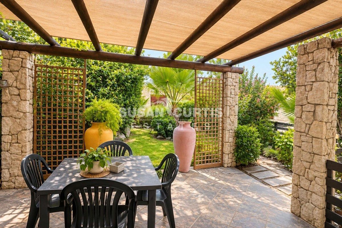 A covered outdoor patio featuring stone-clad pillars, a timber pergola with shade cloth, and tiled flooring, overlooking a landscaped garden.