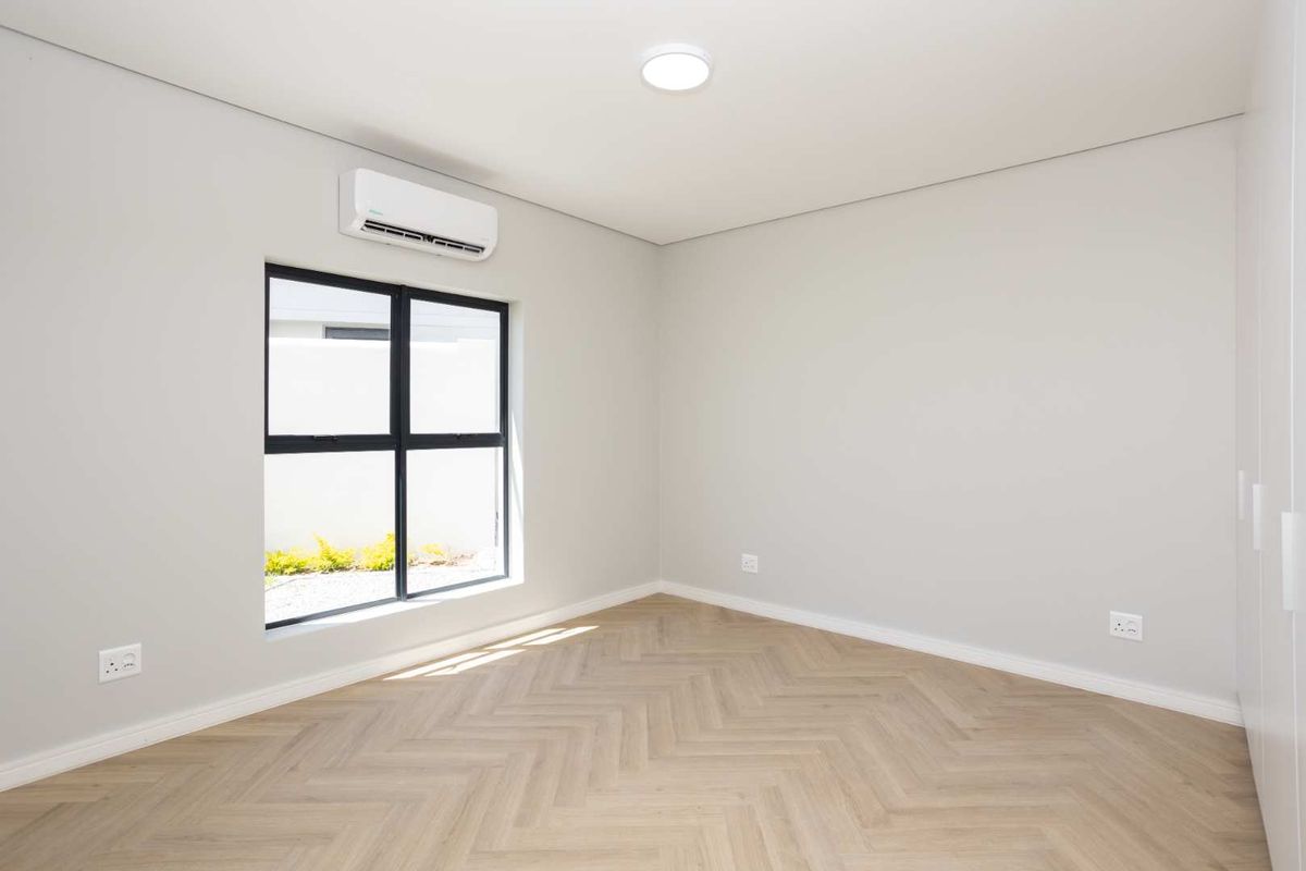 A bright, modern bedroom featuring light wood-look herringbone flooring, neutral walls, a wall-mounted air conditioning unit, and a large window with black frames.