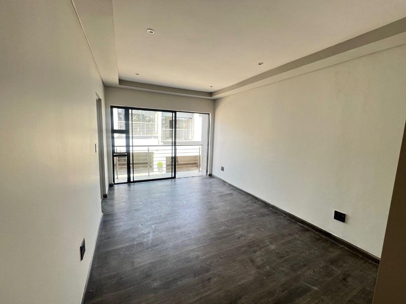A bright, empty bedroom featuring dark wood-look flooring, recessed ceiling lighting, and a large sliding glass door leading to a private balcony.