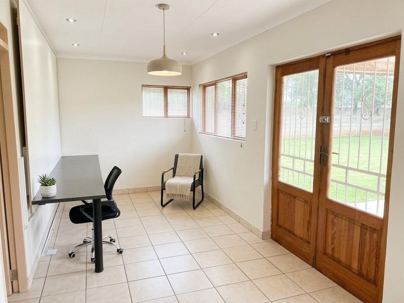 A bright office space featuring tiled flooring, wooden double doors with security gates, and natural light through multiple windows with venetian blinds.