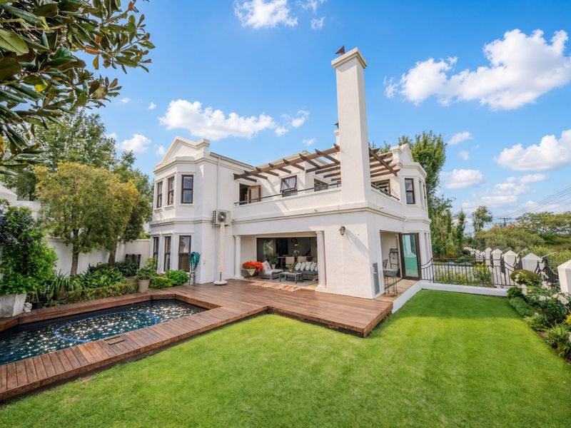 Rear view of a well-maintained two-story home featuring a swimming pool with a wooden deck, a landscaped garden, and an upper-level balcony with a pergola.