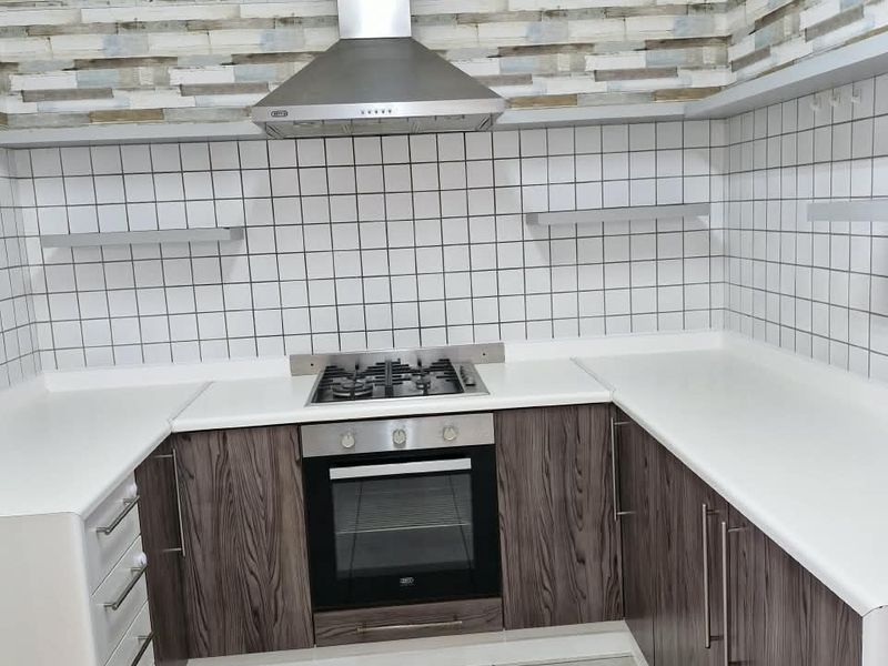 A clean, modern kitchen featuring wood-grain cabinetry, a stainless steel extractor fan, built-in oven, gas hob, and white tiled splashback.