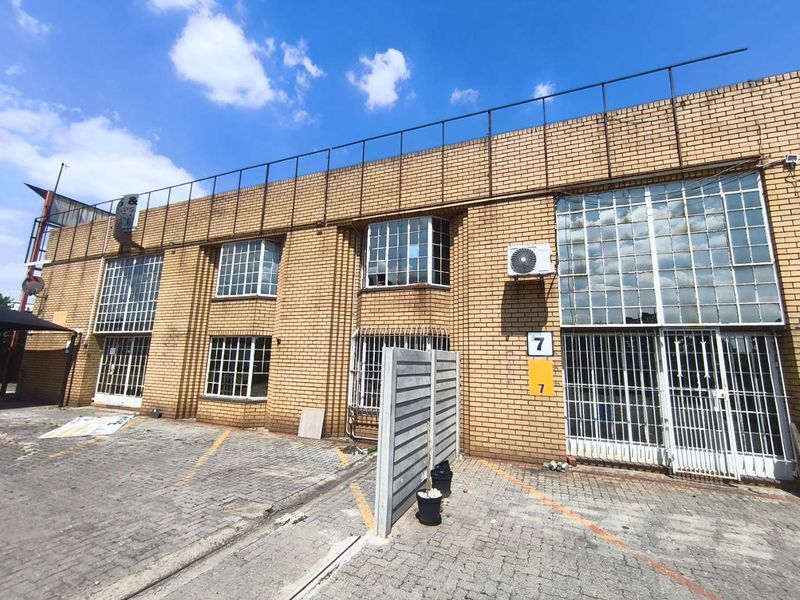 Front elevation of an industrial brick building featuring steel-framed windows, security bars, and a paved parking area.