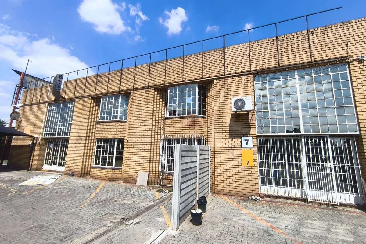 Front elevation of an industrial brick building featuring steel-framed windows, security bars, and a paved parking area.