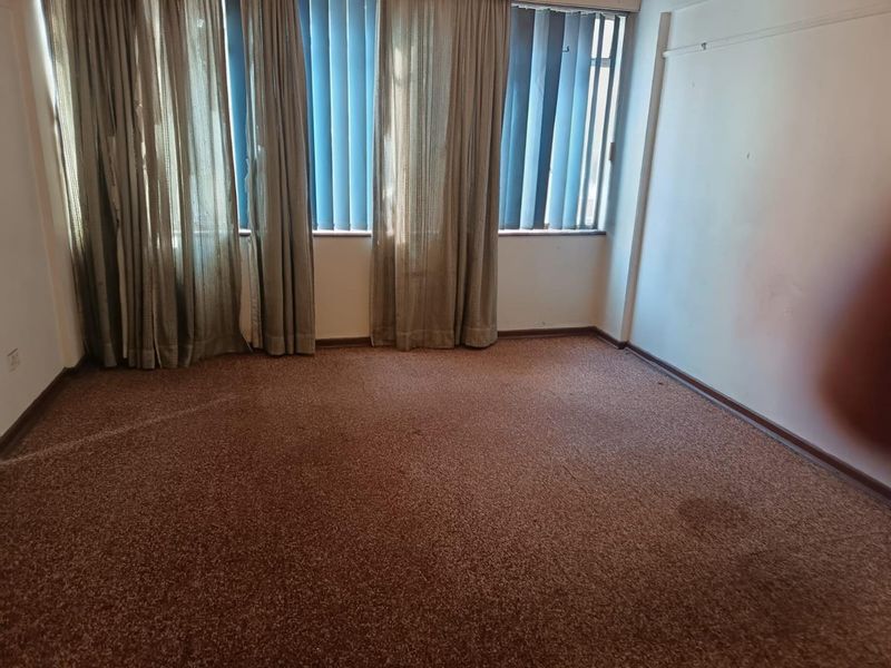 An empty living room featuring wall-to-wall carpeting and vertical window blinds, requiring cosmetic updates to improve its overall condition.