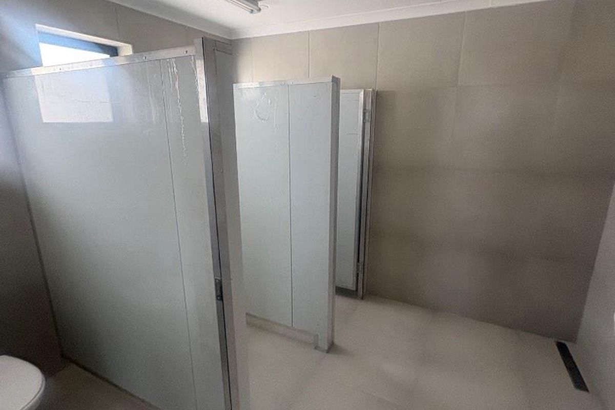 A functional office restroom featuring multiple toilet cubicles with partition walls, tiled flooring, and wall-mounted lighting fixtures.
