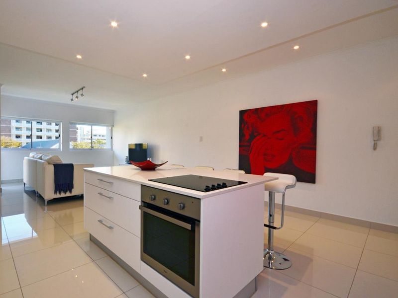 Modern open-plan living area featuring a sleek kitchen island with built-in oven and cooktop, complemented by bright tiled flooring and recessed lighting.