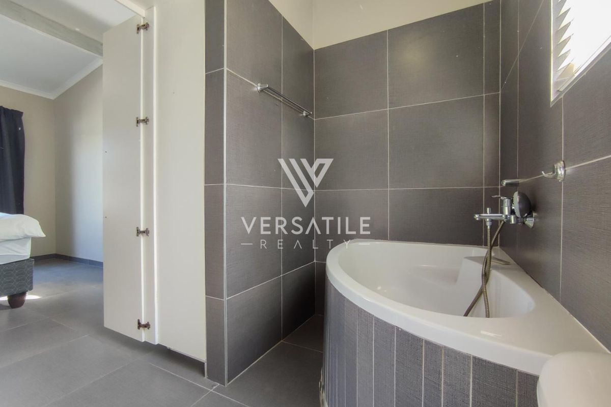A modern bathroom featuring dark grey wall and floor tiling, a built-in corner bathtub with a handheld shower mixer, and a towel rail.