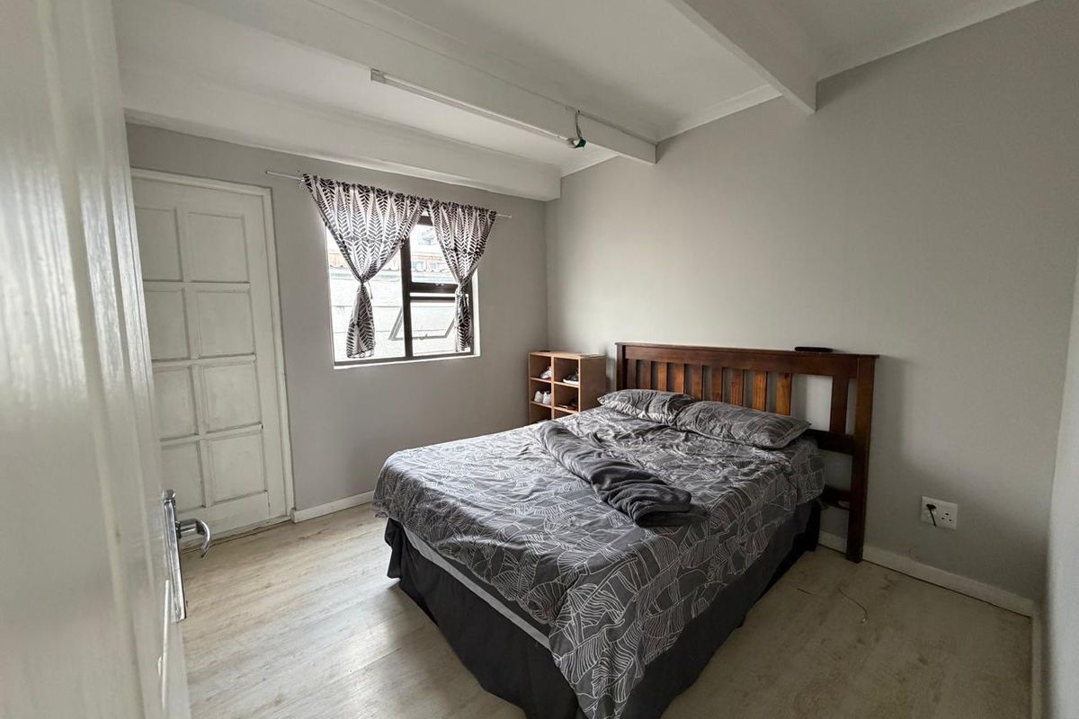 A functional bedroom featuring light-toned laminate flooring, exposed ceiling beams, and a window providing natural light.
