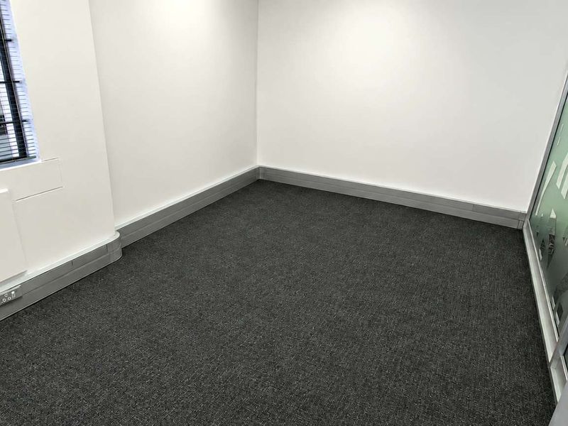 A clean, empty office space featuring dark carpet flooring, a modern glass partition, and an industrial-style window with integrated electrical trunking.