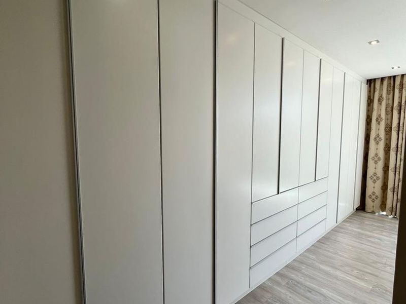 A modern bedroom featuring extensive built-in floor-to-ceiling wardrobes with integrated drawers and sleek laminate flooring.