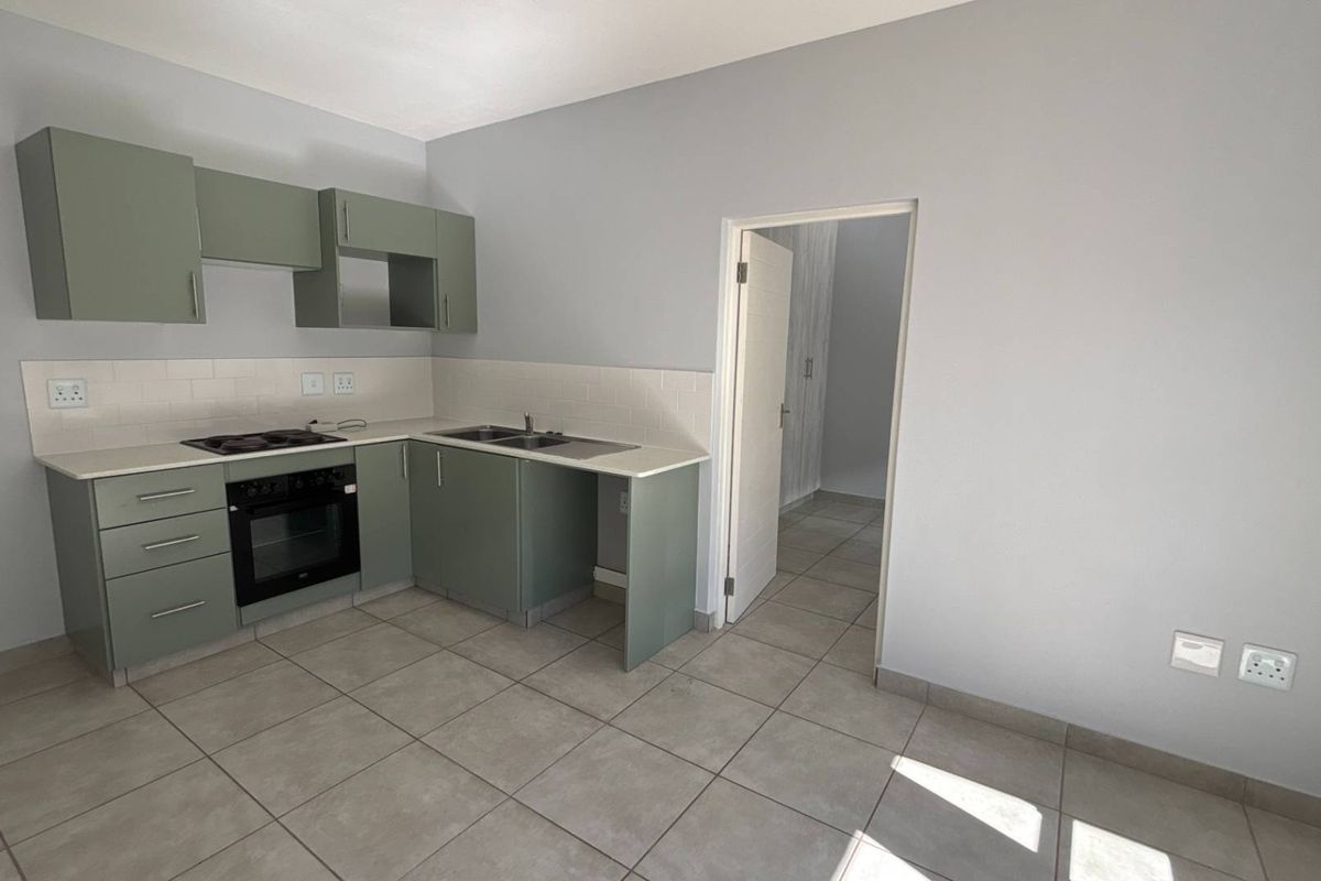 A clean, modern kitchen featuring sage green cabinetry, a built-in oven and hob, tiled backsplash, and neutral floor tiles in an open-plan layout.