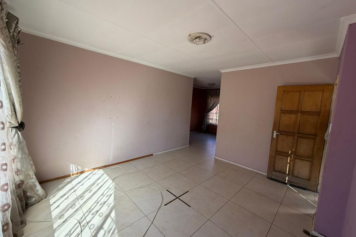 An empty living area featuring tiled flooring, a wooden panel door, and access to an adjoining room, requiring cosmetic updates to refresh the space.