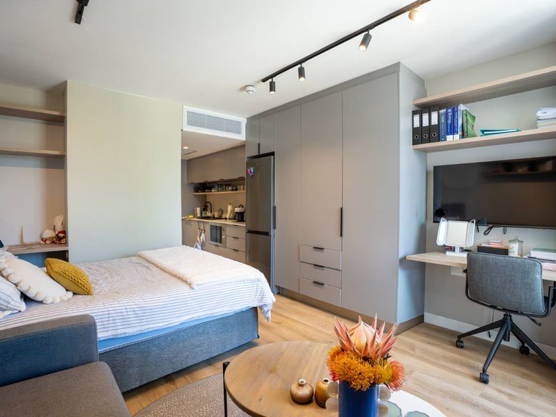 A modern open-plan studio apartment featuring integrated cabinetry, a built-in desk, floating shelving, and contemporary track lighting with light wood-look flooring.