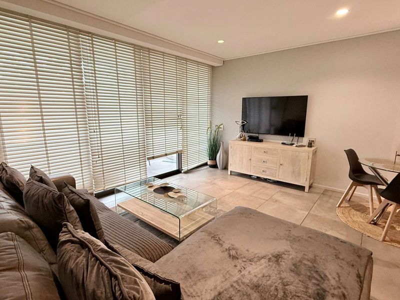 A bright open-plan living and dining area featuring large sliding glass doors with venetian blinds, neutral tiled flooring, and modern recessed downlighting.