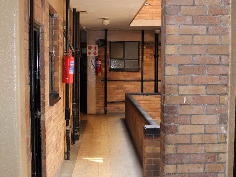 A functional apartment building hallway featuring exposed brick walls, tiled flooring, and visible utility piping with safety equipment.