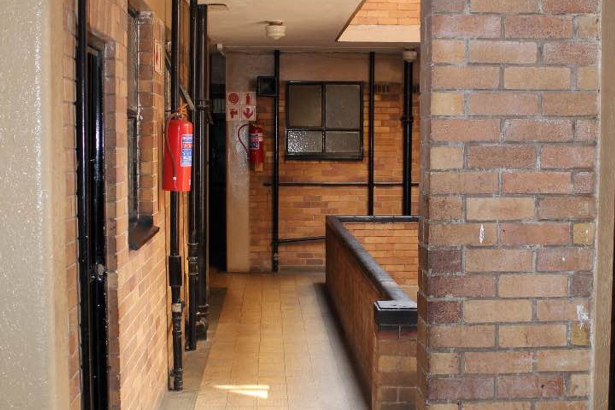 A functional apartment building hallway featuring exposed brick walls, tiled flooring, and visible utility piping with safety equipment.