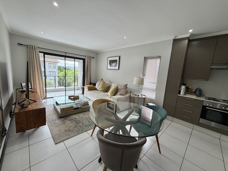 Modern open-plan living area featuring tiled floors, recessed lighting, and a compact kitchen with built-in appliances, leading to a private balcony.