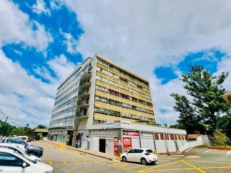 Exterior view of Paige Place, a multi-story apartment building featuring balconies and ground-level commercial space with adjacent paved parking.