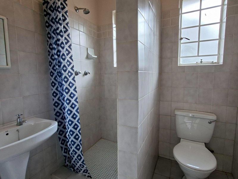A functional bathroom featuring a pedestal basin, a shower enclosure with a curtain, and a standard toilet, finished with neutral wall and floor tiles.