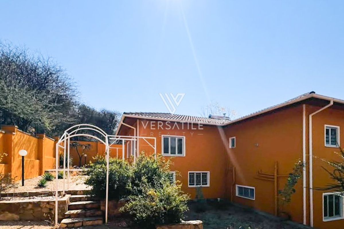 Rear view of the property featuring a stone retaining wall, metal garden arch, and solar water heating system installed on the tiled roof.