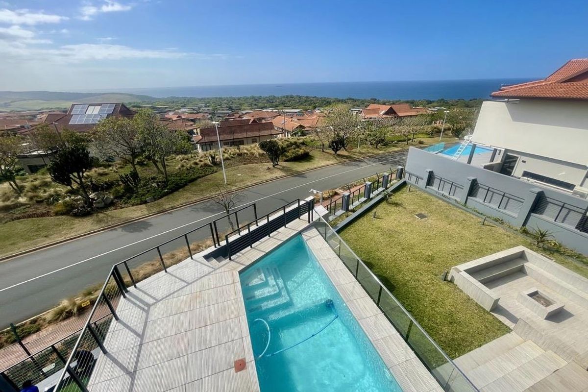 Elevated aerial view of a modern property featuring a swimming pool, glass balustrades, paved patio area, and a landscaped garden with ocean views.