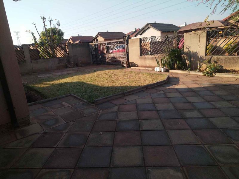 A rear exterior view featuring a paved patio area, a small lawn, and secure boundary walls with metal fencing and a gate.