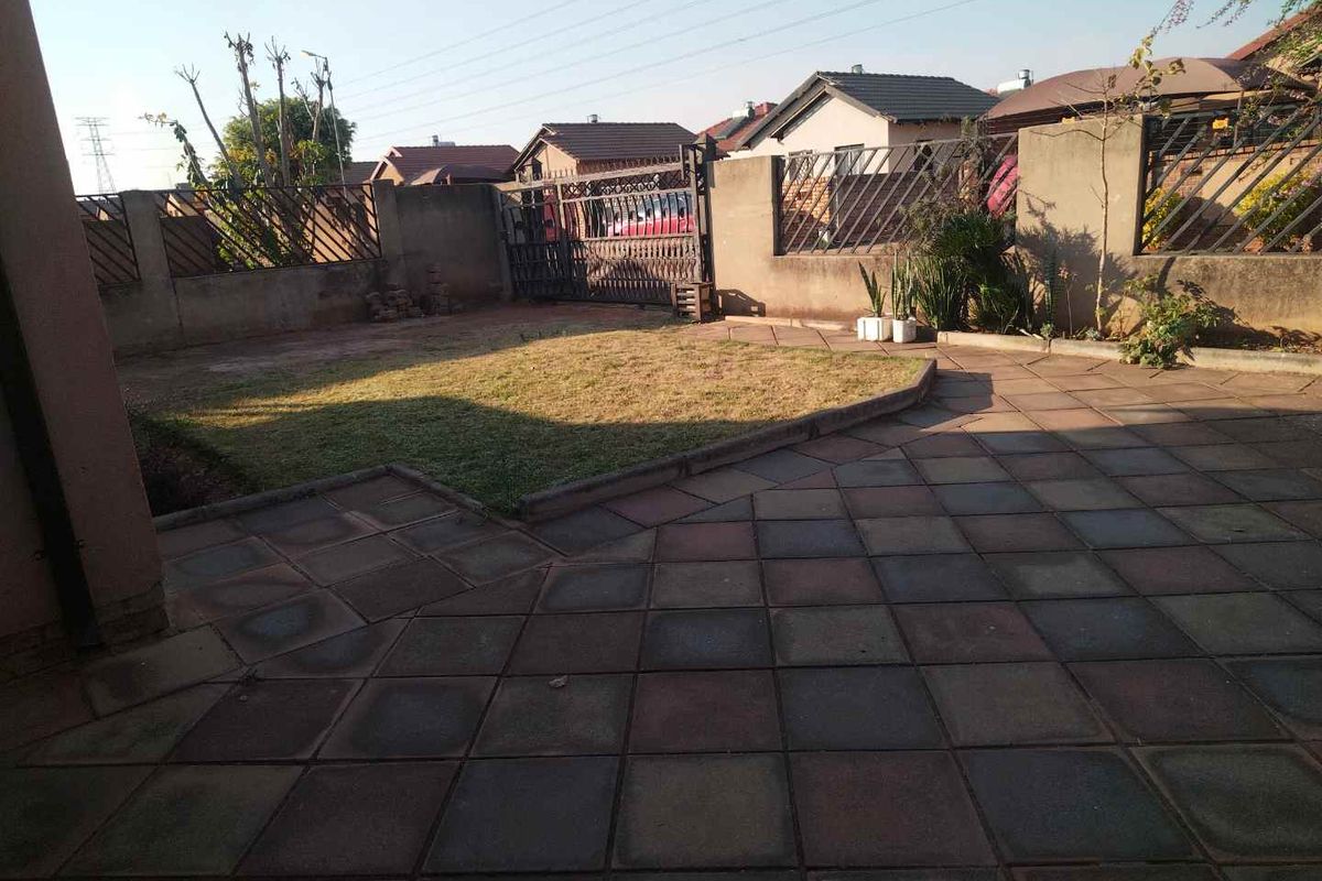A rear exterior view featuring a paved patio area, a small lawn, and secure boundary walls with metal fencing and a gate.
