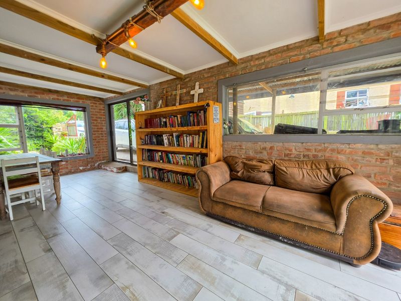 A rustic living area featuring exposed brick walls, timber ceiling beams, and light-toned tiled flooring with large windows and sliding doors providing natural light.