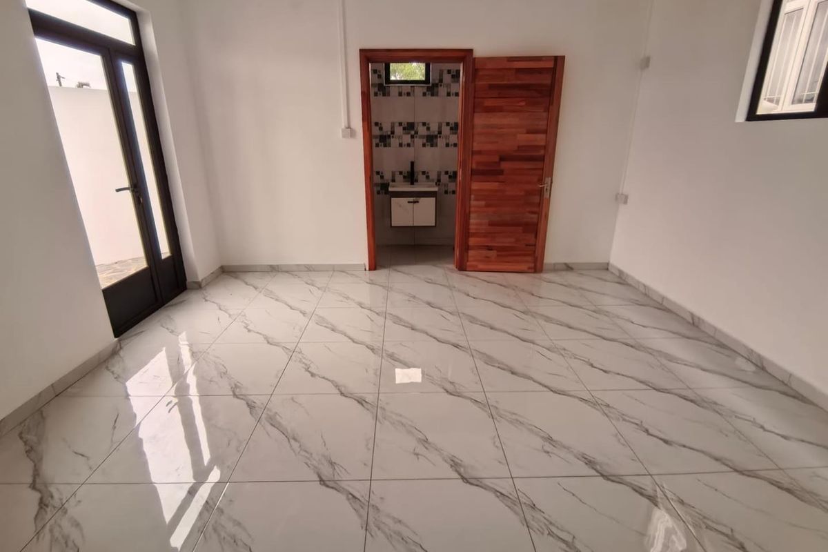 A bright, empty bedroom featuring polished marble-look floor tiles, a wooden door, and an en-suite bathroom with modern vanity.