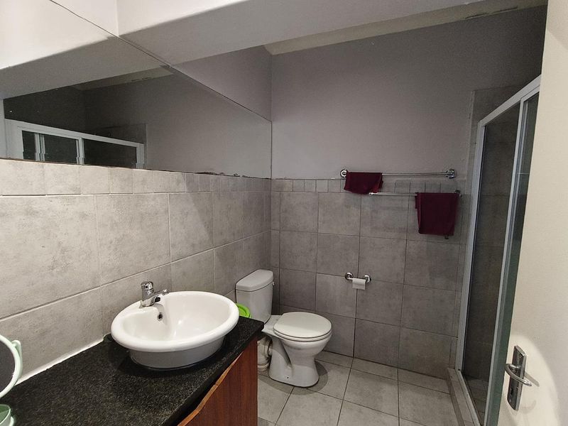 A functional bathroom featuring a vanity with granite countertop, ceramic basin, toilet, and a glass-enclosed shower with neutral wall and floor tiling.