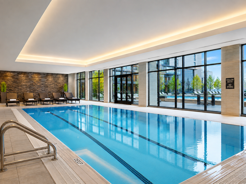 A modern indoor swimming pool area featuring a stone accent wall, recessed ceiling lighting, and expansive floor-to-ceiling windows overlooking the outdoor terrace.