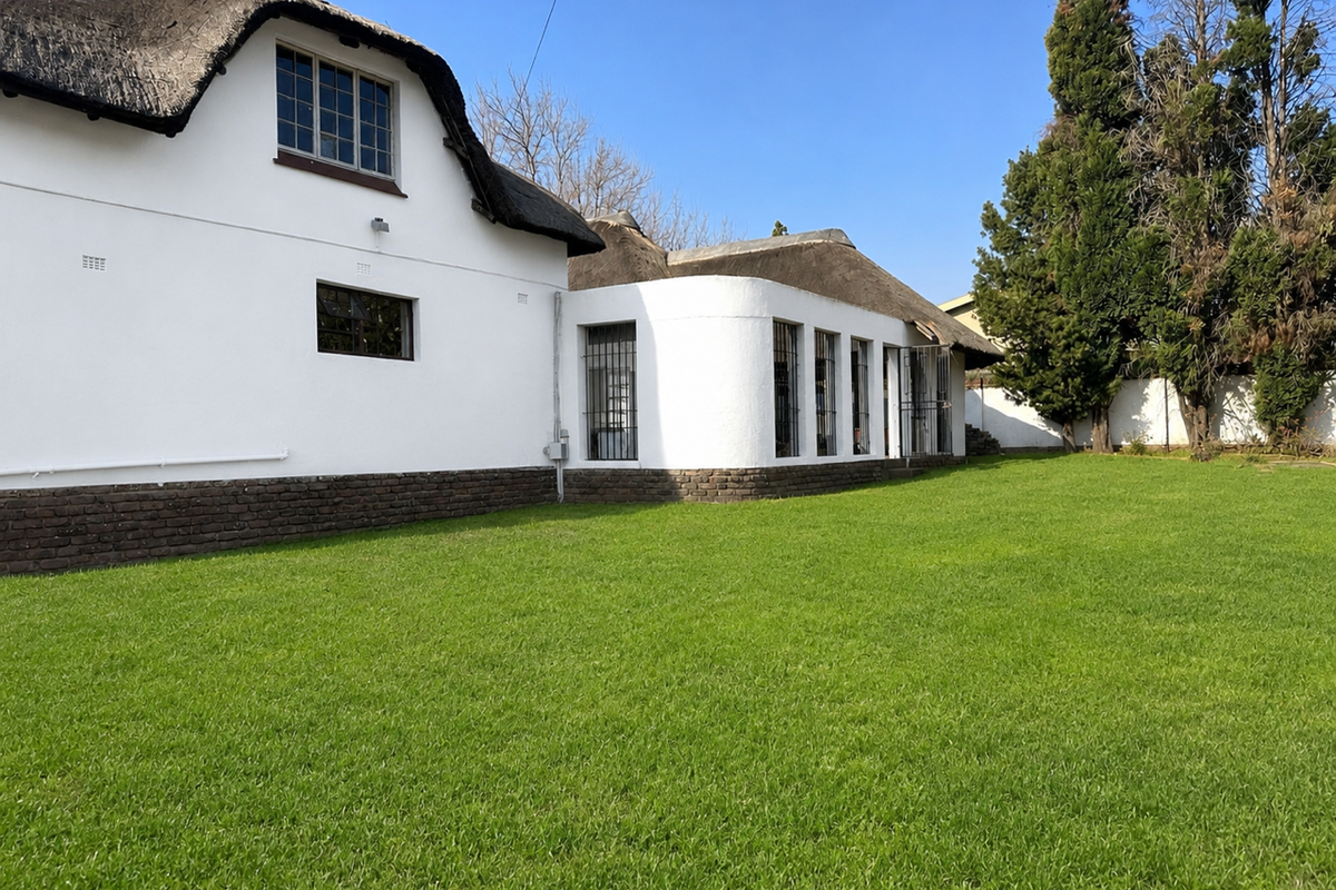 Rear view of a white-walled house featuring a traditional thatch roof, brick base, and a well-maintained green lawn.