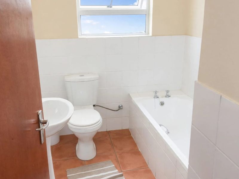 A functional bathroom featuring a pedestal basin, toilet, and bathtub with white wall tiling and terracotta floor tiles.
