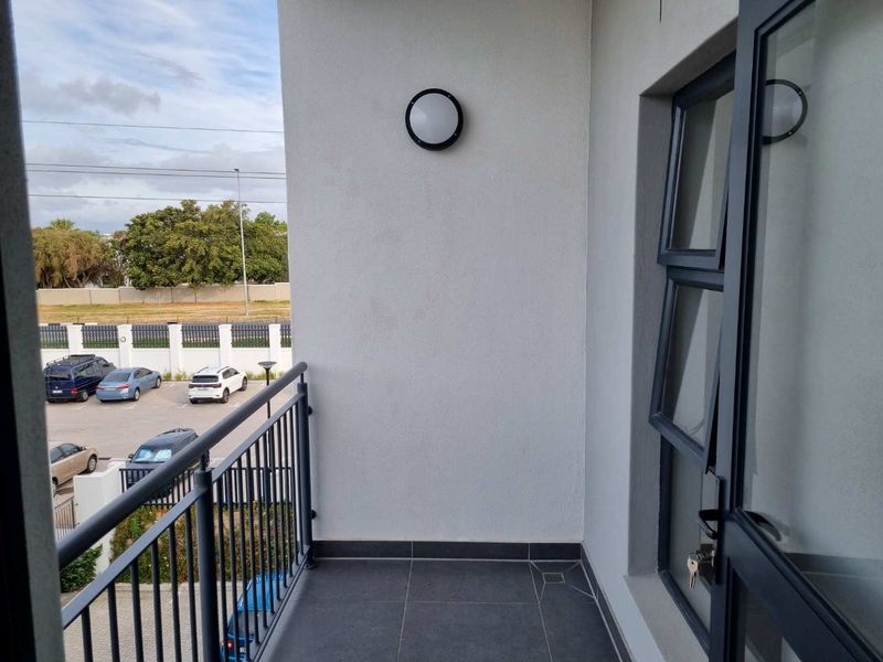 A clean, modern balcony featuring dark tiled flooring, a metal balustrade, and an exterior wall light, overlooking a parking area.