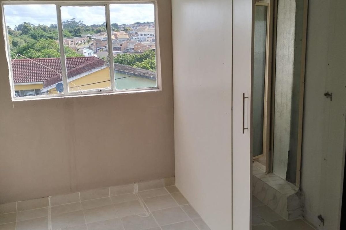 An empty bedroom featuring tiled flooring, a built-in wardrobe, and an en-suite bathroom, with unfinished wall finishes and a view of the surrounding neighbourhood.