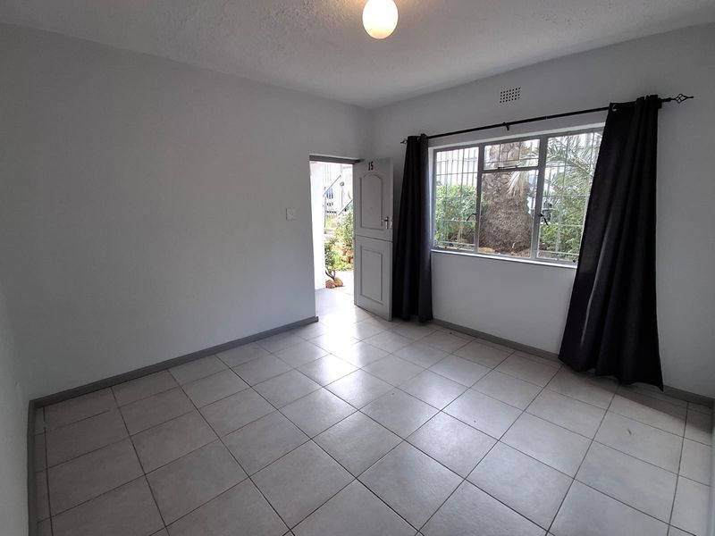 An empty living room featuring neutral tiled flooring, a large window with security bars, and direct access to the exterior entrance.