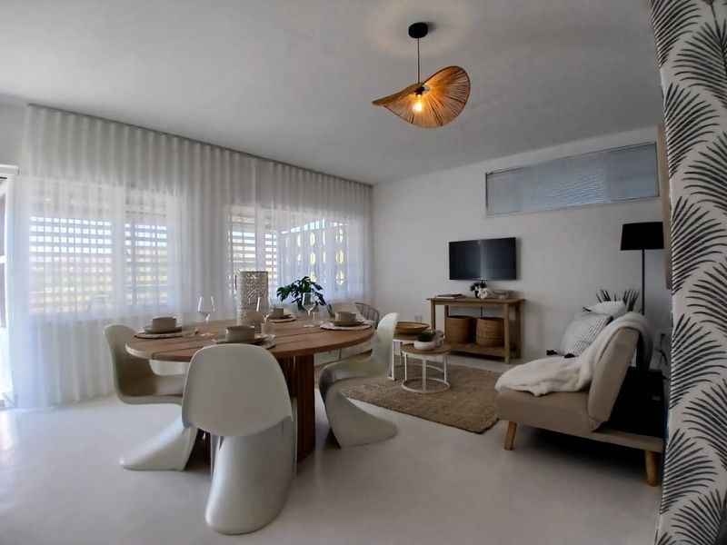 A bright and airy living and dining area featuring neutral tiled floors, modern pendant lighting, and large windows dressed with sheer curtains.