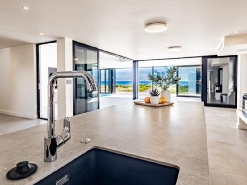 Modern open-plan living area featuring a sleek kitchen island with undermount sink, contemporary fireplace, and expansive sliding doors opening to a coastal view.