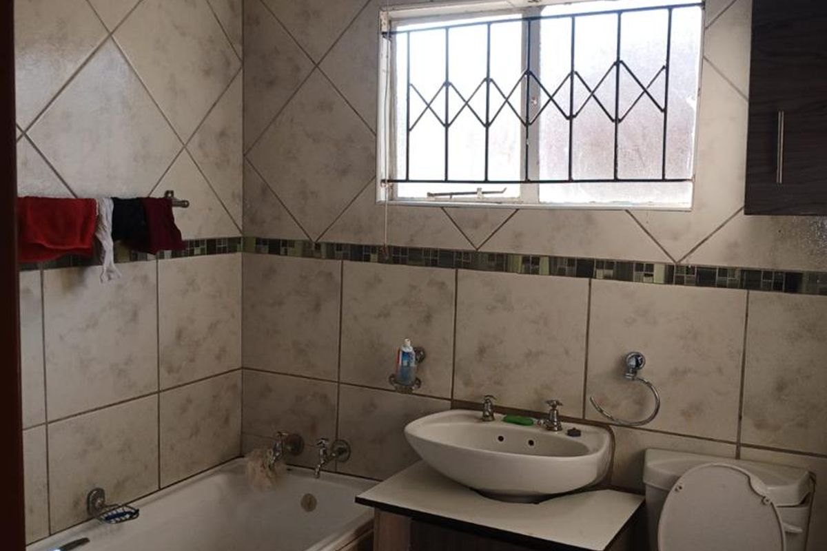 A functional bathroom featuring a built-in bathtub, vanity cabinet with basin, and toilet, finished with tiled walls and security bars on the window.