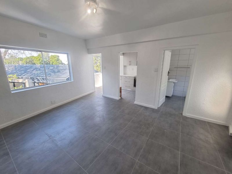 An empty, well-lit living area featuring modern grey floor tiles, white walls, and direct access to the kitchen and bathroom.