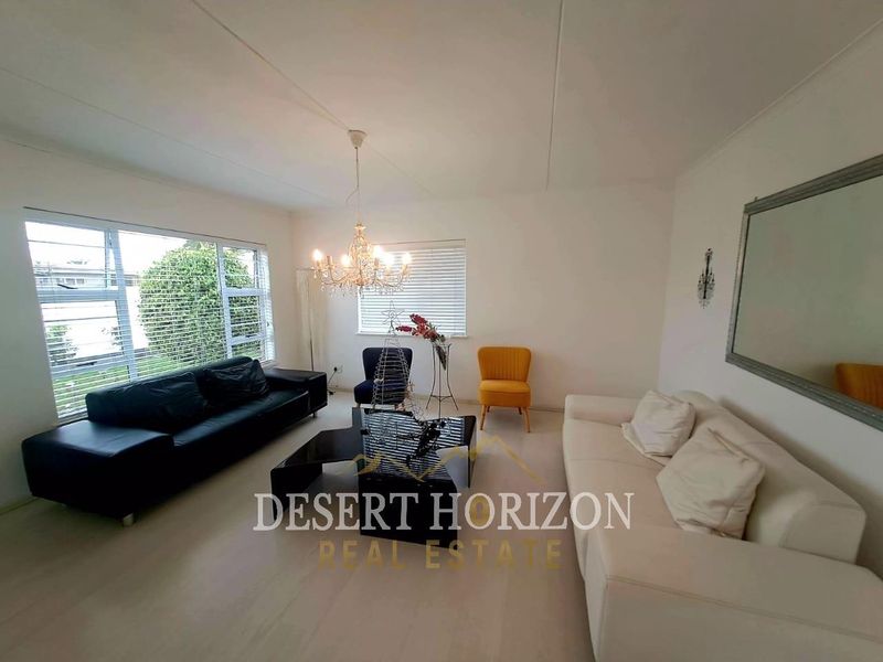 A bright and spacious living room featuring large windows with venetian blinds and a central chandelier, offering a clean and neutral living space.