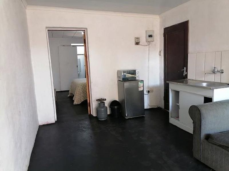 Open plan living area featuring a basic kitchenette with a stainless steel sink and tiled splashback, leading into an adjacent bedroom space.
