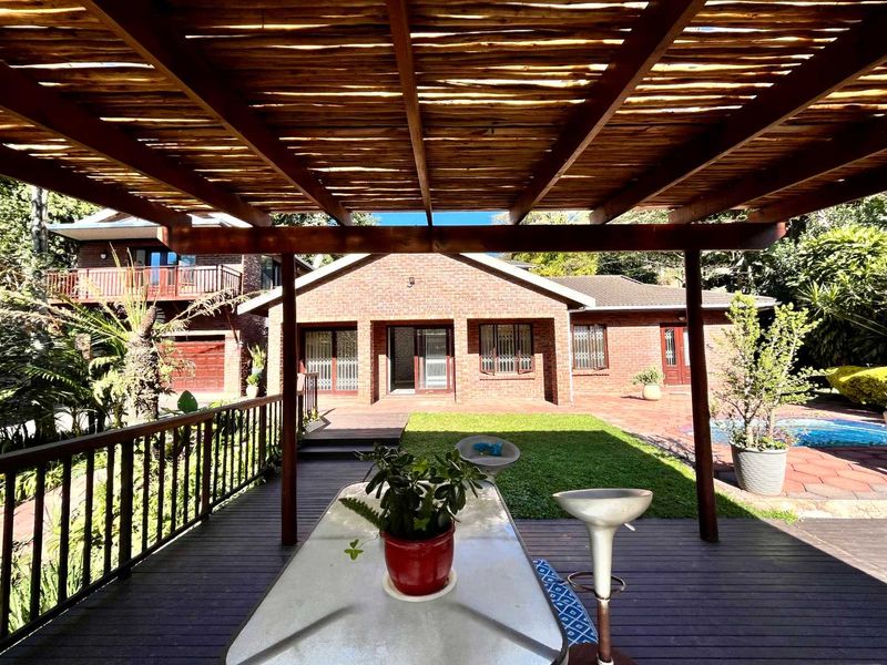 Spacious covered timber deck patio featuring a rustic pergola, overlooking a well-maintained garden and swimming pool area with brick exterior finishes.