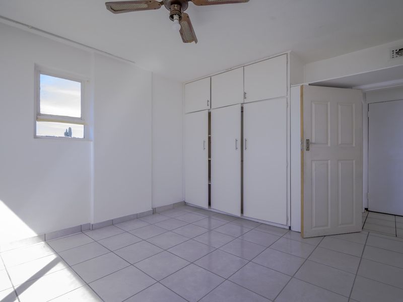 A bright, empty bedroom featuring white tiled flooring, built-in cupboards, and a ceiling fan, offering a clean and neutral space ready for personalization.