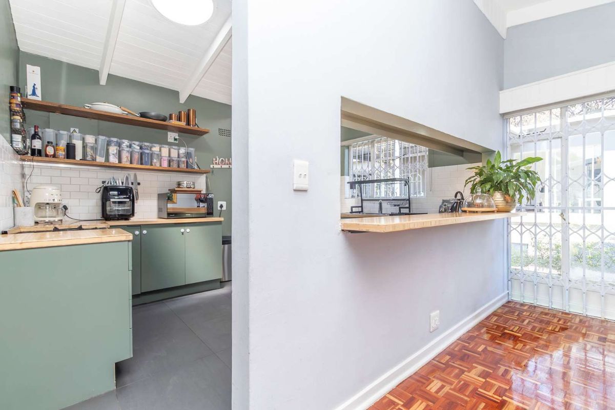 A functional kitchen featuring sage green cabinetry, timber countertops, subway tile backsplash, and open shelving, connected to an adjacent living area via a serving hatch.