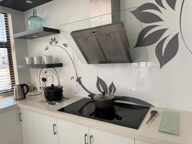 Modern kitchen area featuring a sleek induction hob, stainless steel extractor fan, stone countertops, and decorative glass splashback with floating shelving.